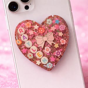 May include: A pink heart-shaped phone grip with the name "RAYLEN" spelled out in colorful letters. The grip is decorated with a pink bow, flowers, stars, and glitter. The phone grip is attached to a white phone.
