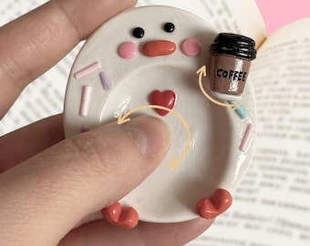 May include: A small, round, white ceramic trinket dish featuring a cartoon face with black eyes, pink cheeks, and an orange beak. The dish is decorated with pastel sprinkles, a central red heart, and a small brown coffee cup labeled COFFEE.
