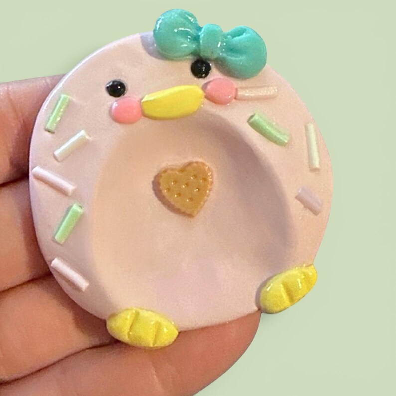 May include: A round, light pink clay trinket dish shaped like a duck face. It features a bright teal bow on top, black eyes, pink cheeks, a yellow beak, and two yellow feet. The surface is decorated with small, light green and white sprinkles.