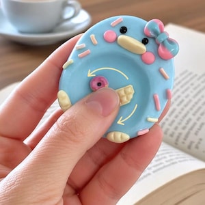 May include: A light blue, oval-shaped fidget toy featuring a cartoon duck face with a pink and blue bow. The surface is decorated with small, raised sprinkles and includes a central rotating element with a small donut and waffle graphic.