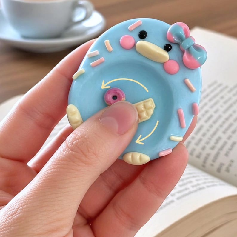 May include: A light blue, oval-shaped fidget toy featuring a cartoon duck face with a pink and blue bow. The surface is decorated with small, raised sprinkles and includes a central rotating element with a small donut and waffle graphic.