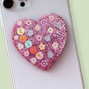 May include: A pink glitter heart phone grip decorated with small flowers and colorful circular accents. The letters "E", "M", "I", and "L" are visible on the circular accents. The phone grip is attached to a white phone.