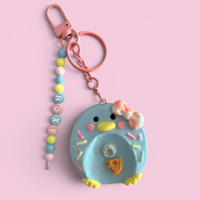 May include: A light blue penguin-shaped keychain with a pink bow, yellow feet, and colorful sprinkles. It features a dangling strand of beads spelling ROXY in white letters, attached to a pink metal lobster clasp and a circular ring.