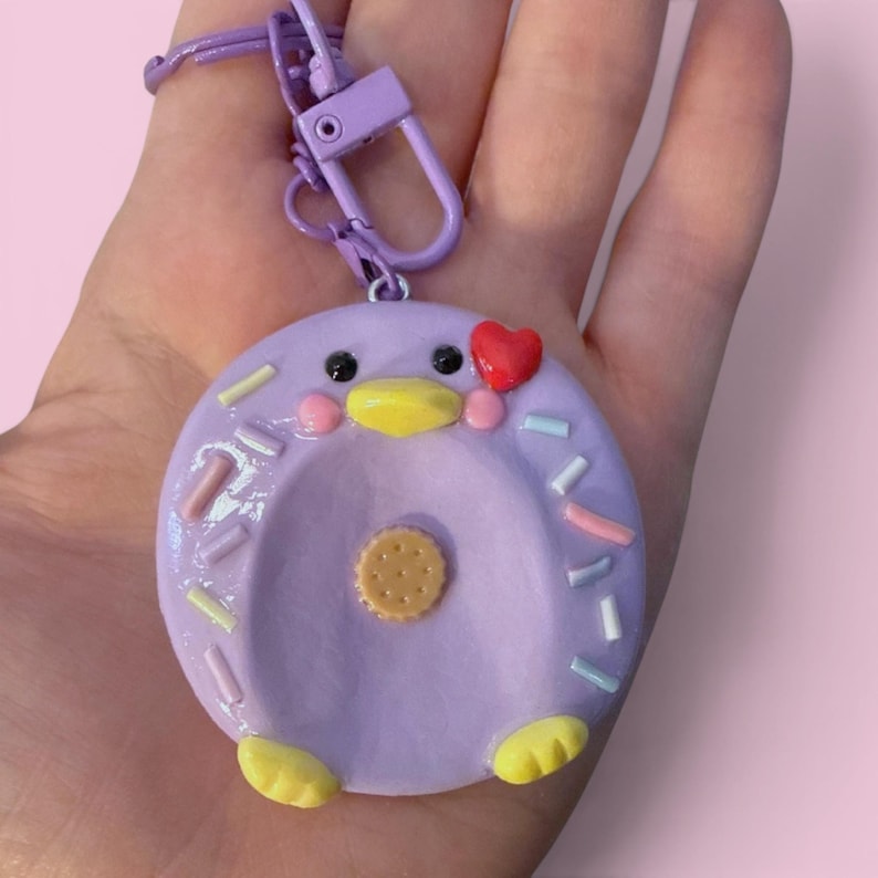 May include: A purple circular keychain charm shaped like a stylized bird with a yellow beak, yellow feet, black eyes, and a small red heart accent. The charm features a light brown circular cookie detail in the center and pastel-colored sprinkles.