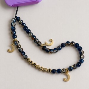 May include: A beaded phone charm with dark blue and clear beads. Gold-colored crescent moon charms dangle from the beaded strand. The name "CAMILLE" is spelled out in gold-colored letter beads.