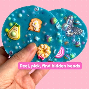 May include: Two round, blue slime toys with embedded colorful beads and charms. The charms include a smiling avocado, a piece of toast with an egg, a butterfly, and a donut. The text "Peel, pick, find hidden beads" is displayed on a pink background.