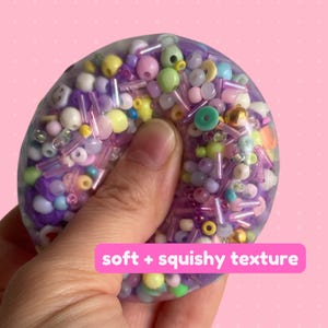 May include: A circular, transparent, squishy sensory toy filled with a variety of colorful beads, including purple, yellow, green, and white spheres and cylindrical tubes. The text soft + squishy texture is printed on a pink rectangular overlay.