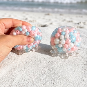 May include: Two clear, jellyfish-shaped stress balls filled with pink, blue, and white beads. One is being squeezed by a hand. The balls have clear, tentacle-like bases and are resting on white sand near the ocean.