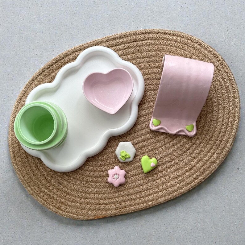 May include: A collection of ceramic items on a woven mat. Includes a white, scalloped tray with a heart-shaped dish and a green cup. Also, a pink, curved stand and several small, decorative pieces in pastel colors.