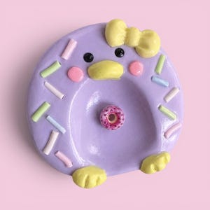 May include: A circular lavender ceramic trinket dish shaped like a duck, featuring a yellow bow, a yellow beak, black eyes, and pink cheeks. The surface is decorated with pastel-colored sprinkles and a small, centered pink donut-shaped accent.