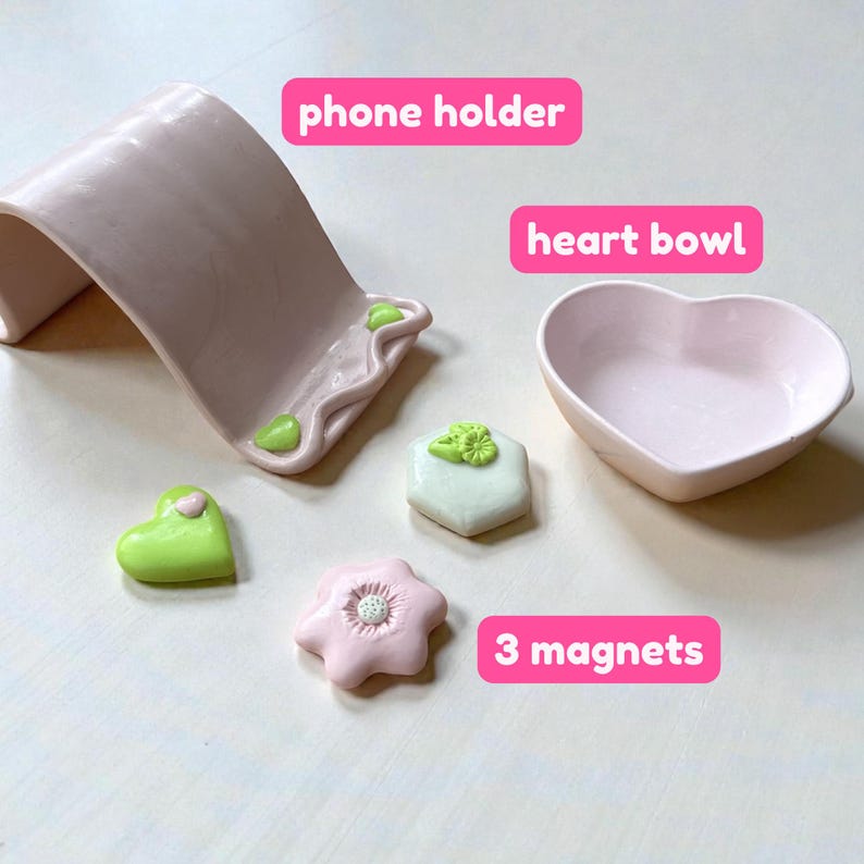 May include: A light pink ceramic phone holder, heart-shaped bowl, and three decorative magnets. The magnets include a green heart, a white hexagon, and a pink flower. The phone holder has green accents.