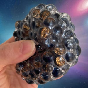 May include: A squishy sensory stress ball featuring a clear, flexible outer mesh casing filled with numerous small, round, translucent spheres in shades of dark brown, black, and amber, creating a textured, bubbly surface appearance.