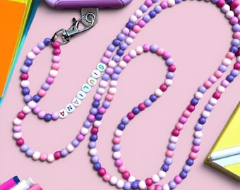 May include: A phone lanyard made of round beads in shades of pink, purple, and lavender. The lanyard features a silver-toned metal lobster clasp attached to a light purple phone case and includes white letter beads spelling GIULIANA.