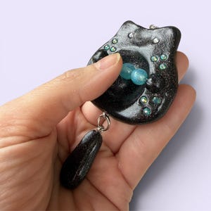 May include: A black, glittery, rounded pendant featuring a central indentation with two light blue translucent beads. The surface is decorated with small, iridescent, circular crystals. A teardrop-shaped black glittery charm hangs from a silver ring.
