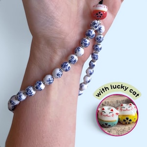 May include: A beaded bracelet featuring white ceramic beads with blue floral patterns, separated by small silver-toned spacers. The bracelet includes a red bead shaped like a cat and an inset showing two additional lucky cat beads.
