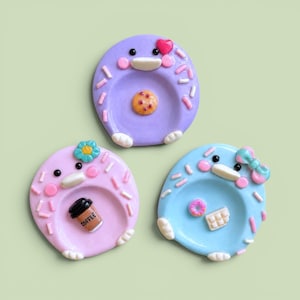 May include: Three round ceramic trinket dishes in purple, light pink, and light blue. Each dish features a cartoon face with pink cheeks, white feet, and colorful sprinkles. The purple dish has a cookie, the pink has a coffee cup, and the blue has a donut and chocolate bar.