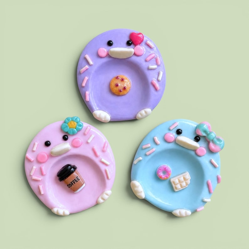 May include: Three round ceramic trinket dishes in purple, light pink, and light blue. Each dish features a cartoon face with pink cheeks, white feet, and colorful sprinkles. The purple dish has a cookie, the pink has a coffee cup, and the blue has a donut and chocolate bar.