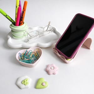 May include: A collection of desk accessories including a phone stand, a cloud-shaped tray, a heart-shaped dish, and decorative shapes. The phone stand is pink with a purple phone. The tray holds glasses and a pen holder with colorful markers.