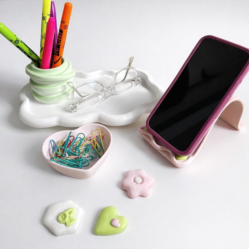 May include: A collection of desk accessories including a phone stand, a cloud-shaped tray, a heart-shaped dish, and decorative shapes. The phone stand is pink with a purple phone. The tray holds glasses and a pen holder with colorful markers.