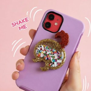 May include: A lavender phone case with a gold glitter dog-shaped shaker charm. The charm contains colorful beads and small white bone-shaped pieces. The text "SHAKE ME" is printed on the case. The phone has a red camera.