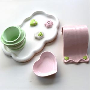 May include: A collection of pastel-colored ceramic items. Includes a white cloud-shaped tray with green and pink decorative accents, a stackable green cup, a pink heart-shaped dish, and a pink paper towel holder with green heart details.