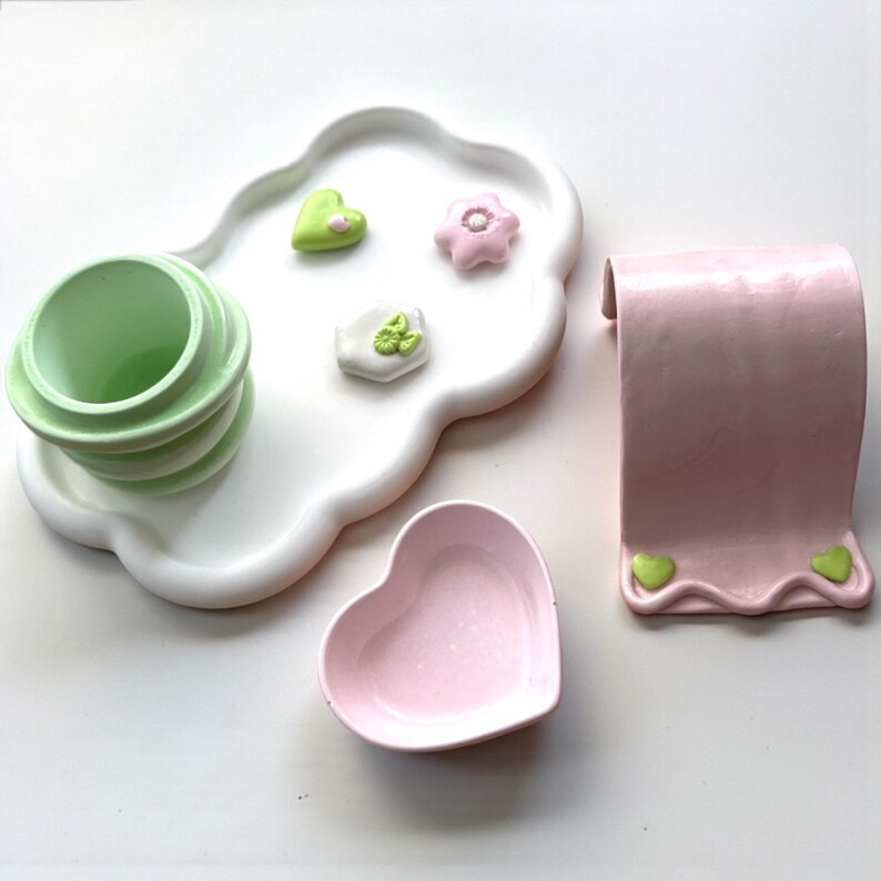 May include: A collection of pastel-colored ceramic items. Includes a white cloud-shaped tray with green and pink decorative accents, a stackable green cup, a pink heart-shaped dish, and a pink paper towel holder with green heart details.