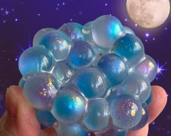 May include: A cluster of translucent, spherical bubbles in shades of blue and purple, forming a stress ball. The bubbles have a shimmering, iridescent effect with gold flecks. The background shows a full moon and a starry night sky.
