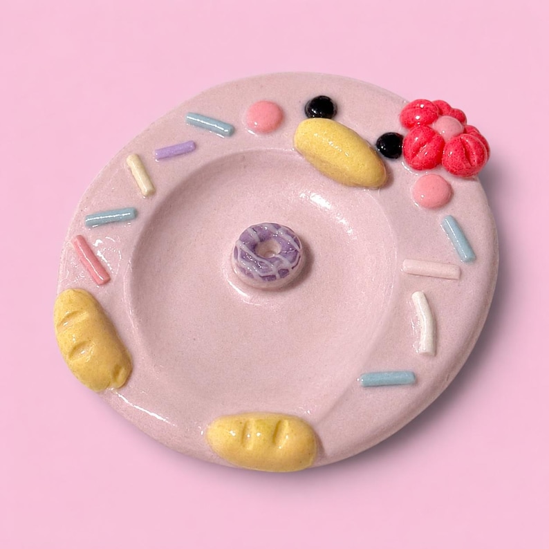 May include: A circular light pink ceramic trinket dish shaped like a donut, featuring a small purple donut in the center, yellow bread-like accents, pastel sprinkles, a pink flower, and two small black dots on a smooth, glossy surface.