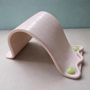 May include: A light pink ceramic sculpture with a curved, arching design. The base features a wavy edge with two small, green heart-shaped accents. The piece has a smooth, matte finish and a minimalist aesthetic.