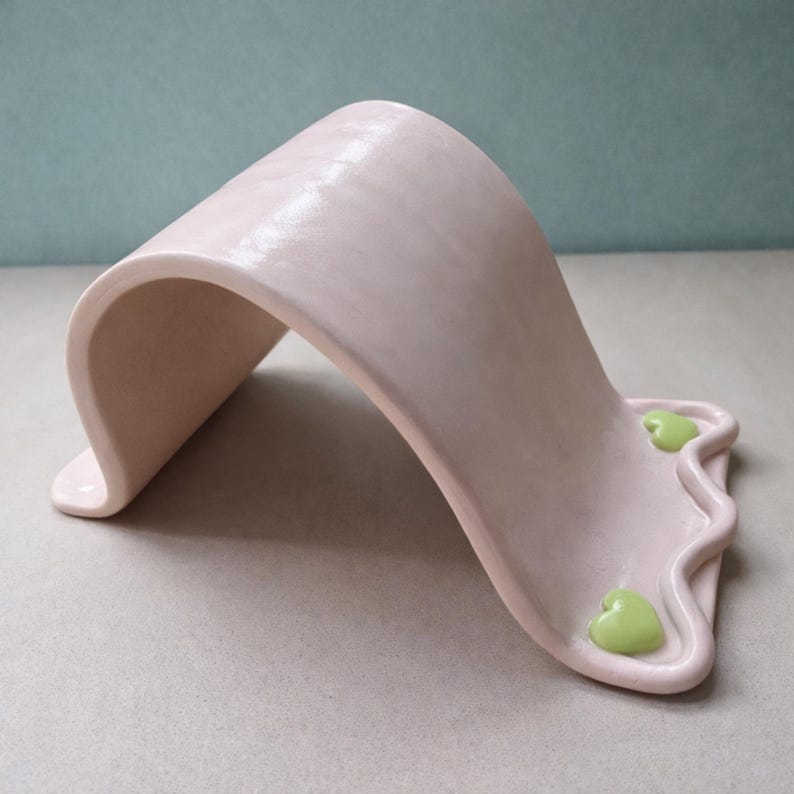 May include: A light pink ceramic sculpture with a curved, arching design. The base features a wavy edge with two small, green heart-shaped accents. The piece has a smooth, matte finish and a minimalist aesthetic.