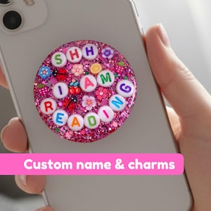 May include: A pink glitter phone grip with the words "SHH I AM READING" spelled out in colorful letter beads. The grip is decorated with small flowers, ladybugs, and fruit charms. The phone grip is attached to a silver phone.