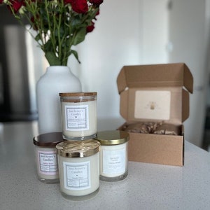 Puede incluir: Cuatro tarros de velas de vidrio con tapas y etiquetas doradas. Las velas están etiquetadas como "Just Breathe Candles" y tienen diferentes aromas. Las velas están dispuestas sobre una encimera blanca.