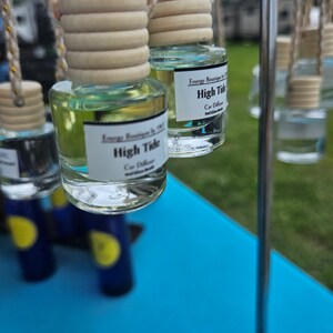 May include: A close-up of three glass car air fresheners hanging from a metal rack. The air fresheners are clear and contain a light blue liquid. The labels on the air fresheners read "High Tide" and "Car Diffuser".