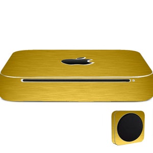 Peut inclure: Un ordinateur doré avec un logo Apple noir sur le dessus. L'ordinateur a une texture métallique brossée et une fente pour un disque. Un carré doré assorti avec un cercle noir est dans le coin inférieur droit.