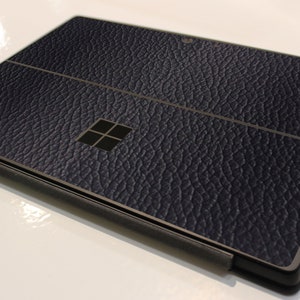 May include: A dark blue laptop with a leather-textured finish, silver trim and the Microsoft logo. The laptop is closed and a black stylus is lying next to it.