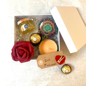 Può includere: Una scatola regalo piena di un piccolo barattolo di miele, una piccola candela, una rosa rossa e caramelle al cioccolato. La scatola è decorata con un'etichetta che dice "Eid Mubarak".