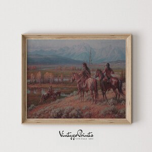 May include: A framed vintage print depicting a landscape with Native American figures on horseback. The artwork shows a mountainous backdrop, a river, and a grassy foreground. The print has a light wood frame and the text "Vintygo Prints Vintage Art".