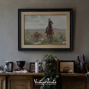 May include: A framed vintage print of a cowboy riding a horse with a herd of cattle in the background. The print is in a black frame and is hanging on a wall above a mantelpiece.