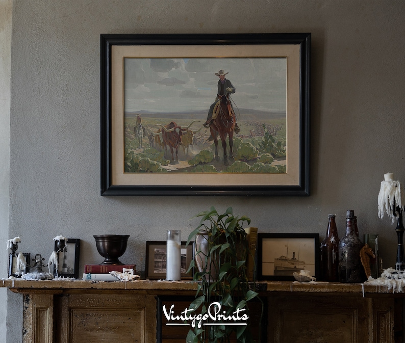 May include: A framed vintage print of a cowboy riding a horse with a herd of cattle in the background. The print is in a black frame and is hanging on a wall above a mantelpiece.