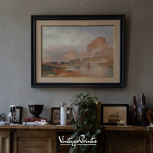 May include: A framed landscape painting of a river scene with mountains in the background, hanging above a fireplace mantel. The mantel is decorated with candles, a vase, books, and a plant.