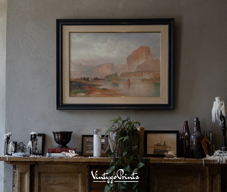 May include: A framed landscape painting of a river scene with mountains in the background, hanging above a fireplace mantel. The mantel is decorated with candles, a vase, books, and a plant.