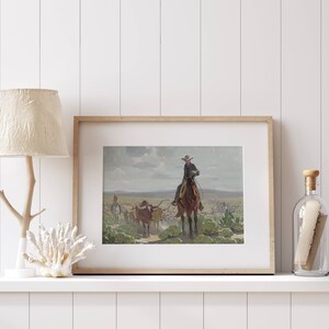 May include: A framed print of a painting depicting a cowboy on horseback leading a herd of cattle across a grassy plain. The sky is cloudy and the scene is set in a rural landscape.