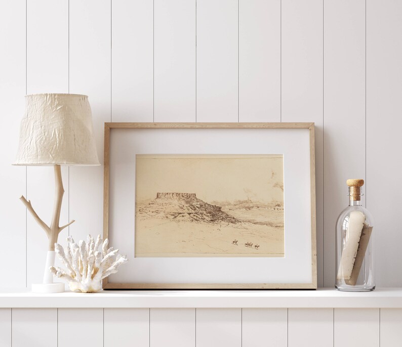 May include: A framed black and white drawing of a landscape with a mountain range in the distance. The drawing is on a white background and is framed in a light wood frame. The frame is sitting on a white shelf with a white lamp and a glass bottle with a cork stopper.