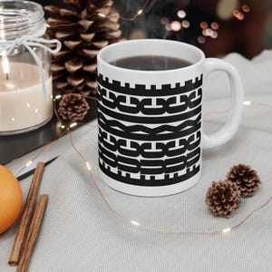May include: White ceramic mug with a black geometric pattern filled with a dark liquid. The mug has a curved handle and is surrounded by decorative elements, including a lit candle, pine cones, and an orange slice.