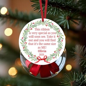 May include: A round white Christmas ornament featuring a green leaf and red berry wreath design. The text reads: This ribbon is very special as you will soon see. Take it out and you will find that it’s the same size as ME! Olivia 2026. A red ribbon hangs from the top.
