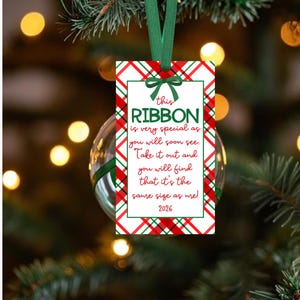 May include: A rectangular white tag with a red and green plaid border, featuring a green ribbon bow at the top. The text reads: this RIBBON is very special as you will soon see. Take it out and you will find that it's the same size as me! 2026.