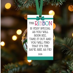 Peut inclure: Une étiquette d'ornement rectangulaire blanche avec un nœud en ruban sarcelle en haut et des bandes sarcelle en haut et en bas. Le texte indique : THIS RIBBON IS VERY SPECIAL AS YOU WILL SOON SEE. TAKE IT OUT AND YOU WILL FIND THAT IT'S THE SAME SIZE AS ME! 2026.