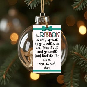 Puede incluir: Una bola de Navidad de cristal transparente con una cinta dorada en su interior, colgada de un hilo dorado. Una etiqueta blanca con detalles turquesa y el texto "this RIBBON is very special as you will soon see. Take it out and you will find that it's the same size as me! 2026."
