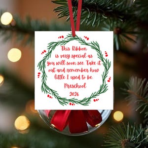 May include: A square white Christmas ornament featuring a green wreath illustration and red text that reads: This Ribbon is very special as you will soon see. Take it out and remember how little I used to be. Preschool 2026. It hangs by a red ribbon.