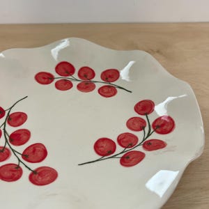 May include: A white ceramic plate with a scalloped edge featuring a hand-painted pattern of three clusters of red berries on thin green stems arranged in a circular formation across the surface of the dish.